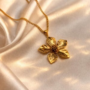 Puede incluir: Un collar dorado con un colgante de flor. El colgante tiene un dise&ntilde;o texturizado y detallado con cinco p&eacute;talos y un centro. La cadena del collar tambi&eacute;n es dorada y descansa sobre una tela sedosa de color crema.