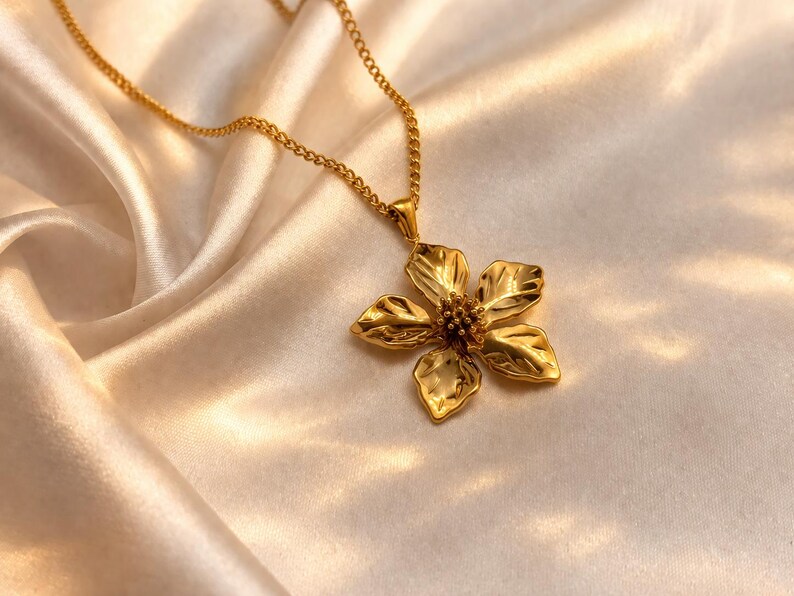 Puede incluir: Un collar dorado con un colgante de flor. El colgante tiene un dise&ntilde;o texturizado y detallado con cinco p&eacute;talos y un centro. La cadena del collar tambi&eacute;n es dorada y descansa sobre una tela sedosa de color crema.