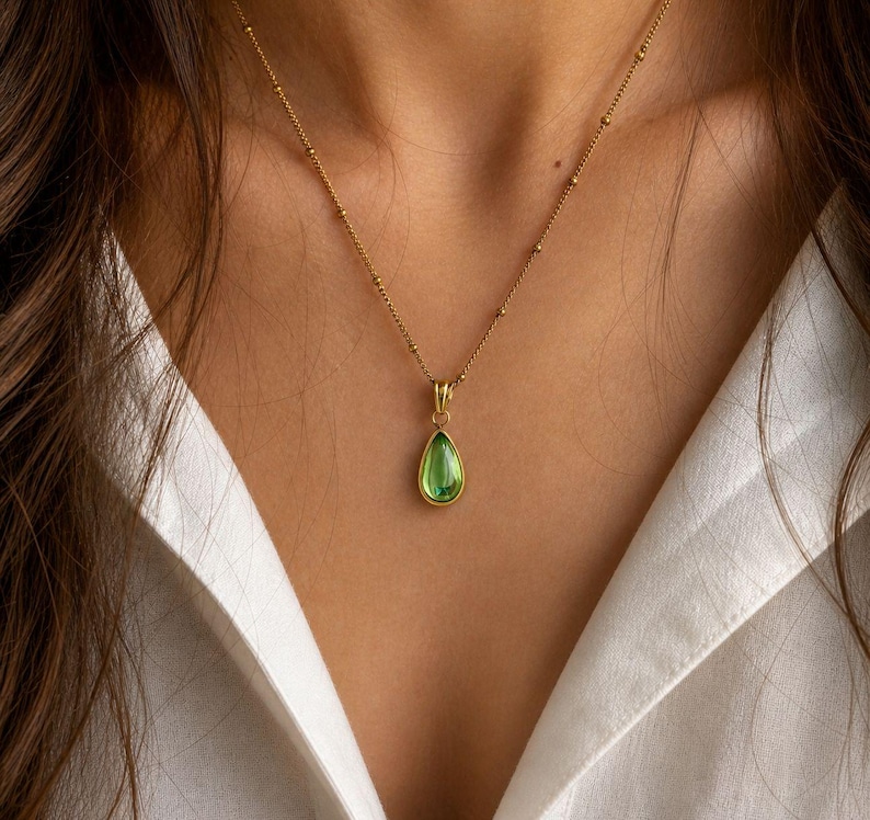 May include: A gold-colored necklace with a teardrop-shaped, green gemstone pendant. The pendant is set in a gold bezel and hangs from a delicate chain with small, evenly spaced beads. The necklace is displayed against a white shirt.