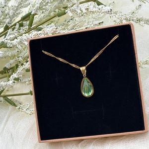 May include: A gold-colored necklace with a teardrop-shaped, light green pendant. The necklace is presented in a black box with a light brown border, set against a backdrop of white flowers.