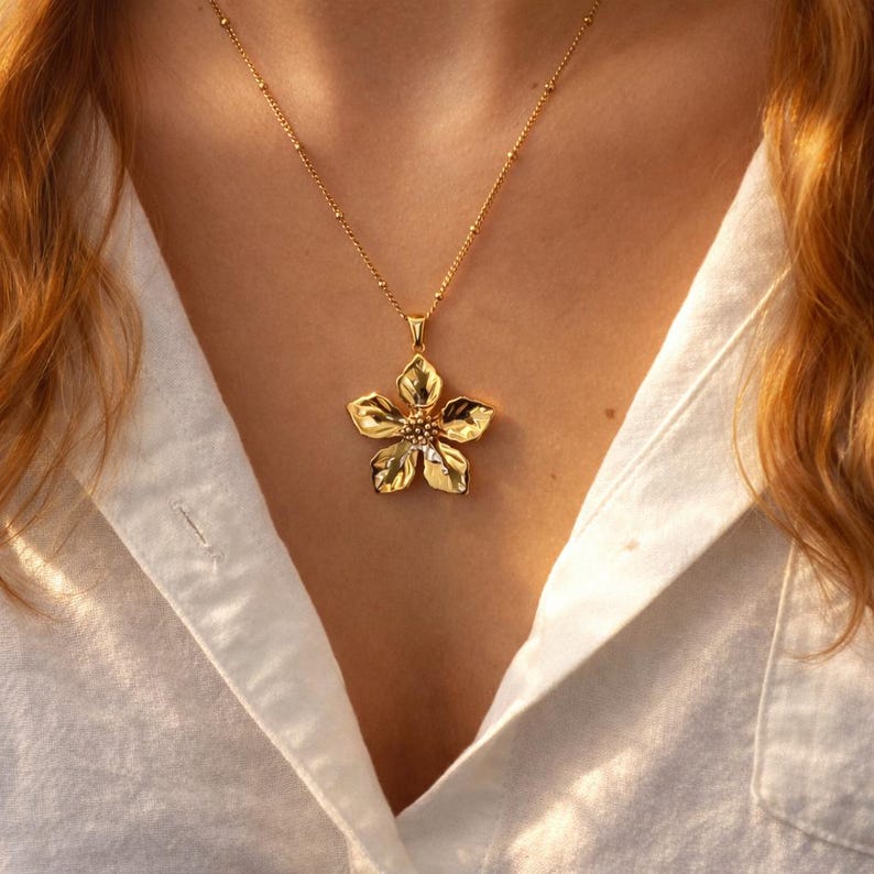 Puede incluir: Un collar dorado con un colgante de flor. El colgante tiene cinco p&eacute;talos y un centro texturizado. El collar se lleva sobre una camisa blanca. La cadena es delicada y tiene peque&ntilde;as cuentas redondas.