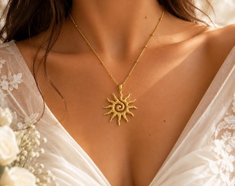 Puede incluir: Un collar de tono dorado con un colgante en forma de sol texturizado que presenta un diseño de espiral central. El colgante cuelga de una delicada cadena dorada que incluye pequeñas cuentas esféricas espaciadas, sobre una prenda blanca.