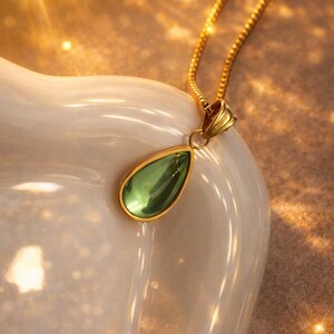 May include: A gold necklace with a teardrop-shaped green pendant. The pendant is framed in gold and hangs from a delicate gold chain. The necklace is displayed on a white, curved surface.
