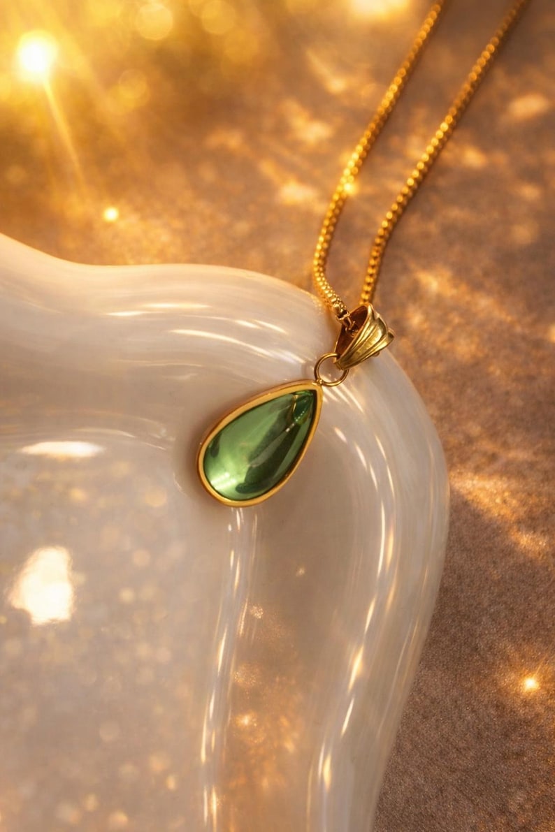 May include: A gold necklace with a teardrop-shaped green pendant. The pendant is framed in gold and hangs from a delicate gold chain. The necklace is displayed on a white, curved surface.