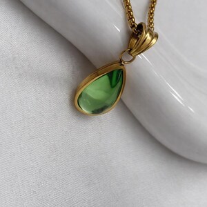 May include: A gold-colored necklace with a teardrop-shaped pendant. The pendant has a light green, transparent stone set in a gold-colored bezel. The chain is a delicate, gold-colored link chain.