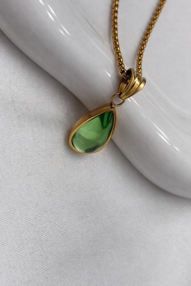May include: A gold-colored necklace with a teardrop-shaped pendant. The pendant has a light green, transparent stone set in a gold-colored bezel. The chain is a delicate, gold-colored link chain.