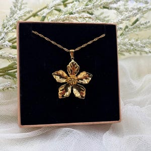Puede incluir: Un collar con colgante de flor dorada presentado en una caja negra. El colgante es una flor detallada de cinco p&eacute;talos con un centro texturizado. La cadena del collar es delicada y retorcida. La caja est&aacute; dentro de una caja marr&oacute;n, con flores blancas en el fondo.