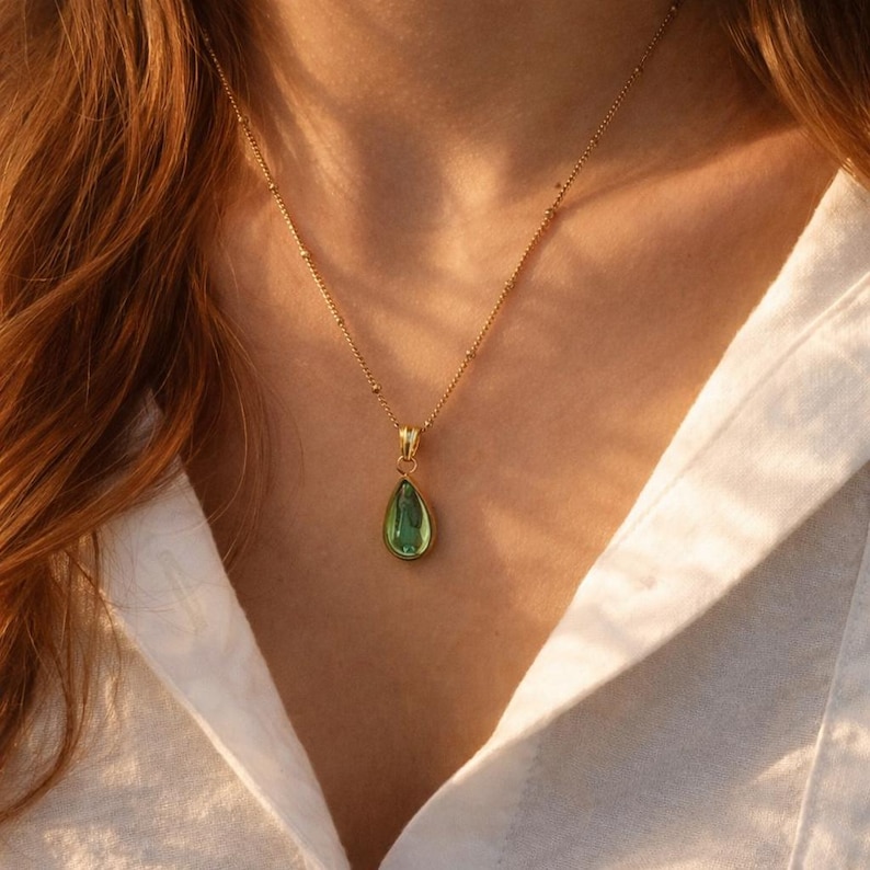 May include: A gold-colored necklace with a teardrop-shaped green pendant. The pendant is set in a gold frame and hangs from a delicate chain. The necklace is worn against a white shirt.