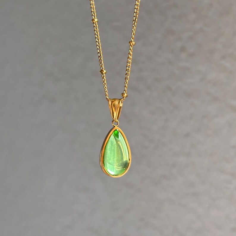 May include: A gold-colored necklace with a teardrop-shaped pendant. The pendant features a light green stone set in a gold frame. The chain is delicate, with small gold beads. The background is a solid gray.