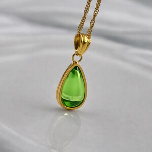 May include: A gold-colored necklace featuring a pear-shaped, bright green pendant. The pendant is encased in a gold frame and hangs from a delicate gold chain. The gemstone reflects light, creating a sparkling effect. The jewelry is displayed on a white surface.