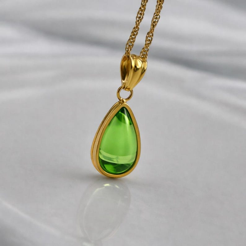May include: A gold-colored necklace featuring a pear-shaped, bright green pendant. The pendant is encased in a gold frame and hangs from a delicate gold chain. The gemstone reflects light, creating a sparkling effect. The jewelry is displayed on a white surface.
