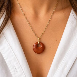 May include: A gold-colored chain necklace with a circular, reddish-brown pendant. The pendant has a hole in the center and a polished finish. The necklace is worn against a white shirt.