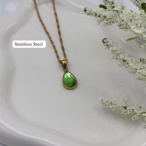 May include: A gold-colored stainless steel necklace with a green teardrop-shaped pendant. The pendant has a gold-colored frame and a vibrant green center. The text "Stainless Steel" is visible.