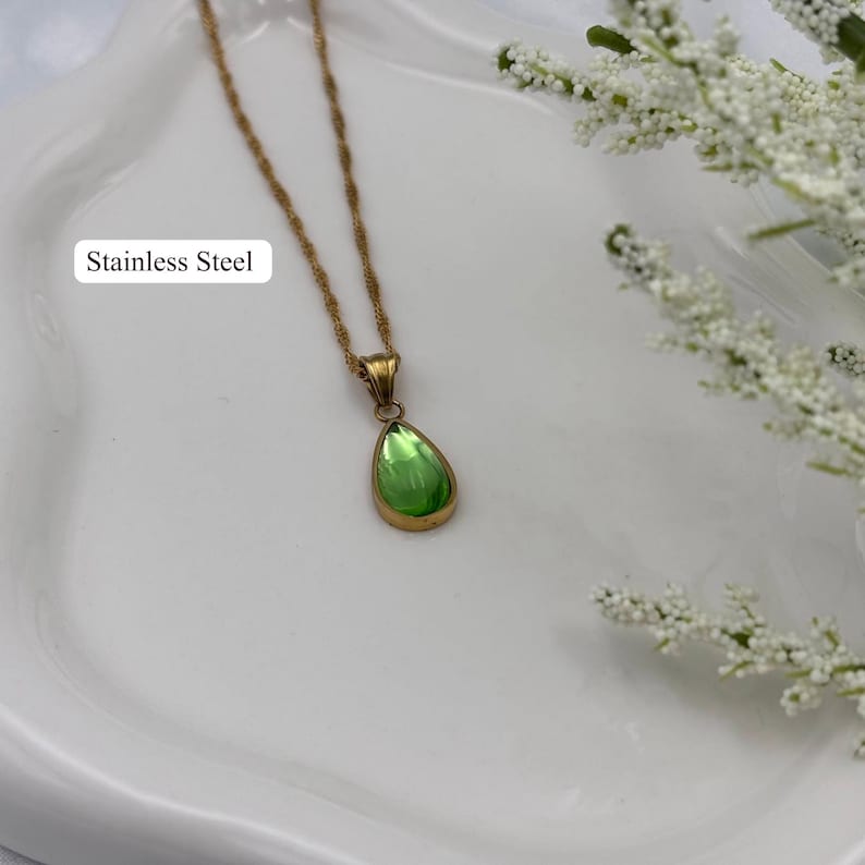 May include: A gold-colored stainless steel necklace with a green teardrop-shaped pendant. The pendant has a gold-colored frame and a vibrant green center. The text "Stainless Steel" is visible.