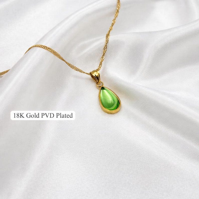 May include: A gold-colored necklace with a teardrop-shaped, light green pendant. The pendant is framed in gold and hangs from a delicate chain. The text "18K Gold PVD Plated" is visible in the image.