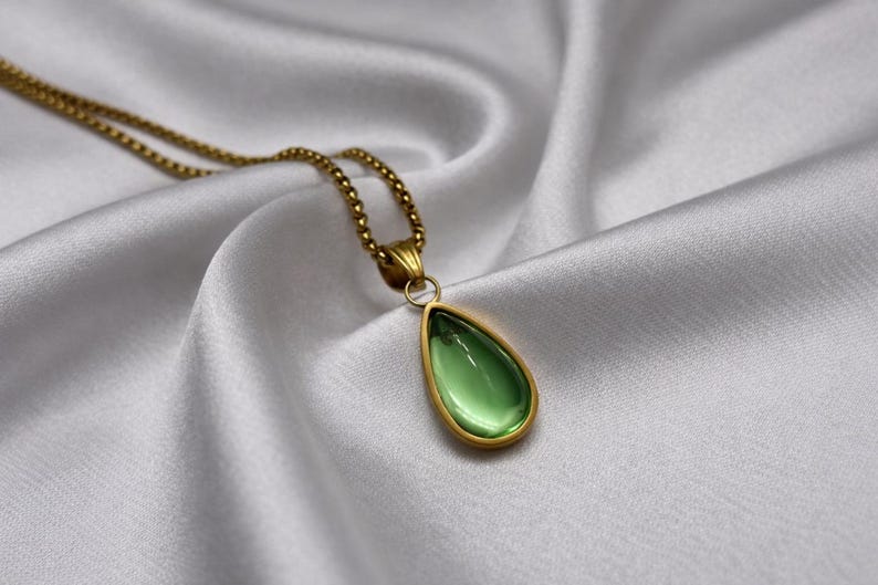 May include: A gold-colored necklace with a teardrop-shaped pendant. The pendant features a light green gemstone set in a gold frame. The necklace rests on a white, silky fabric.