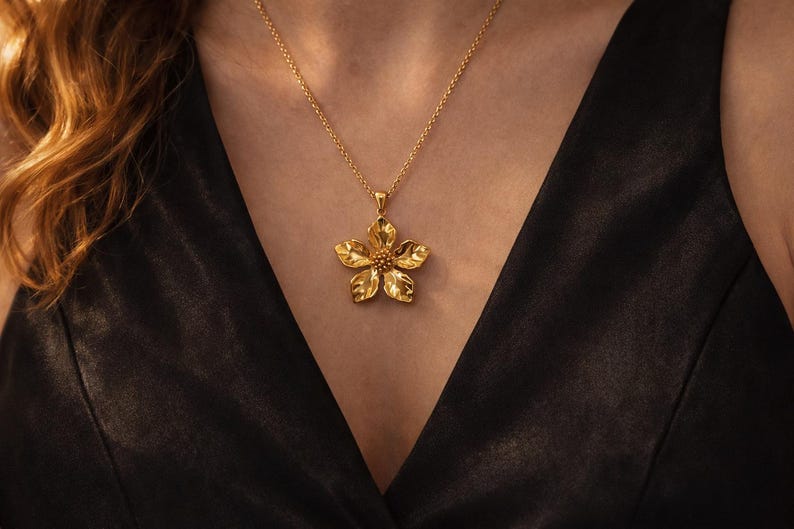 May include: A gold-colored flower pendant necklace. The pendant is a detailed, five-petal flower with a textured surface and a small, round center. The necklace chain is delicate, and the pendant hangs against a black top.