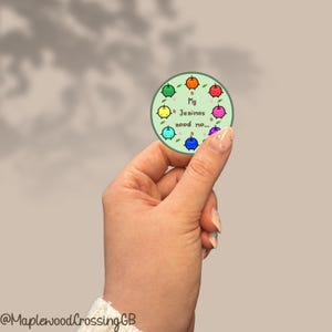 Könnte beinhalten: Ein runder Aufkleber mit hellgrünem Hintergrund, der acht kleine, bunte Cartoon-Figuren in einem Kreis angeordnet zeigt. Der Text in der Mitte lautet "My Junimos need me...". Die Figuren sind orange, rot, rosa, lila, blau, türkis, gelb und grün.