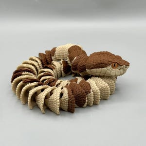 May include: A coiled articulated snake figurine featuring a repeating pattern of textured brown and cream-coloured segments. The snake has a triangular head with detailed scale patterns and prominent orange eyes, designed with flexible joints.
