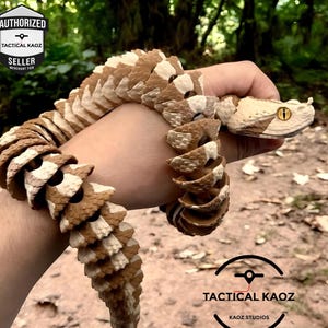 May include: An articulated snake sculpture in brown and cream, held in a hand. The snake's head has a realistic eye and detailed scale patterns. The image includes the text "TACTICAL KAOZ" and "AUTHORIZED SELLER".