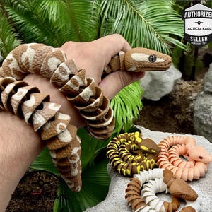 May include: A brown and white articulated snake toy is held in a hand, with the snake's body wrapped around the wrist. Several other coiled snake toys in various colours are also visible, including yellow, orange, and white. The image includes the text "AUTHORIZED TACTICAL KAOZ SELLER".