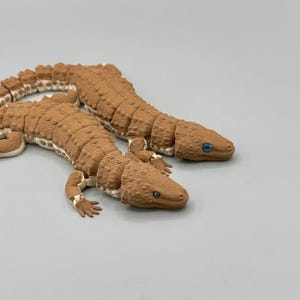 May include: Two articulated lizard figurines with textured brown bodies and cream-coloured underbellies. Each lizard features a segmented design, small limbs with visible toes, and bright blue eyes set into a detailed, scaled head structure.