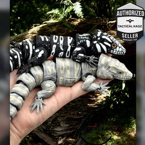 May include: Two articulated lizard figurines with segmented bodies. The top lizard is black with white markings, and the bottom lizard is light grey with tan accents. Both feature detailed scales and eyes. A logo in the corner reads AUTHORIZED TACTICAL KAOZ SELLER.