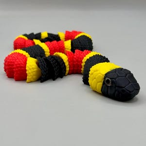 May include: A flexible, articulated toy snake featuring a repeating pattern of vibrant red, bright yellow, and matte black segments. The snake has a textured, scale-like surface throughout its body and a detailed head with visible eye markings.