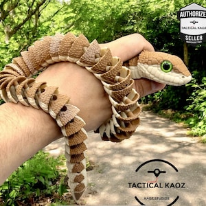 May include: An articulated snake toy in shades of brown and tan, with green eyes, is held in a hand. The snake is made of interlocking segments. The head is tan and brown, with a white underbelly. The background is a blurred outdoor scene. The text "TACTICAL KAOZ" is visible.