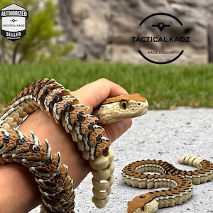 May include: A detailed snake figurine with a tan and brown pattern, held in a hand. The snake's head is tan with yellow eyes. A coiled snake figurine is also on the ground. The text "TACTICAL KAOZ" is visible.