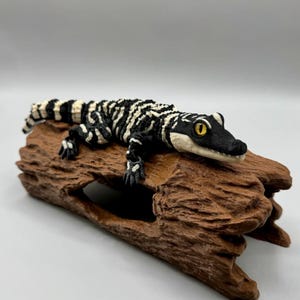 May include: A black and white articulated alligator figurine with bright yellow eyes, resting on a textured brown log. The reptile features a high-contrast striped pattern across its body, head, and tail, with a detailed, realistic skin texture.