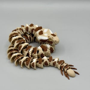 May include: A segmented, articulated toy snake featuring a textured, cream-coloured body with brown patches. The snake is coiled in a circular position, displaying a detailed head with yellow eyes and a distinct, smooth, light-coloured tail tip.