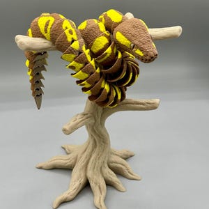 May include: A brown and yellow articulated snake figurine coiled around a light brown textured tree branch stand. The snake features a segmented body with a detailed scale pattern, a pointed tail, and visible eyes, resting on a base shaped like roots.
