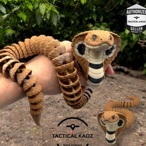 May include: A brown and tan articulated cobra snake toy. The snake has a detailed head with black and white markings and a segmented body. The toy is held in a hand and also shown on the ground. The text "TACTICAL KAOZ" is visible.