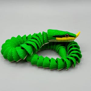 May include: A bright green articulated toy snake with a segmented body, featuring a yellow snout, black eye markings, and brown eyes. The snake is coiled in a circular shape, displaying a textured, scale-like pattern across its entire length.