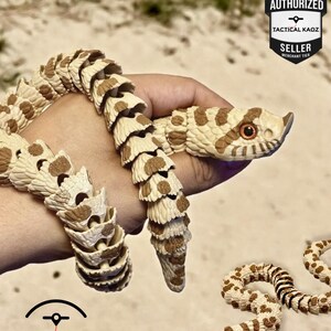 May include: An articulated snake toy in cream and brown, held in a hand. The toy's segmented body and realistic design are visible. The image includes the text "AUTHORIZED TACTICAL KAOZ SELLER".