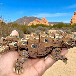 May include: A brown, articulated, 3D-printed horned lizard figurine featuring a segmented body, textured scales, and dark circular markings on its back and limbs. The lizard has a spiky head, a wide tail, and light-coloured claws on its feet.