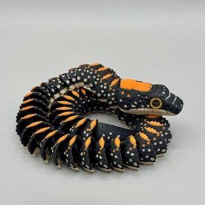 May include: A segmented, articulated toy snake featuring a black body with white spots and bright orange accents along the spine and head. The snake is coiled in a circular position, showcasing a detailed scale texture and prominent yellow eyes.