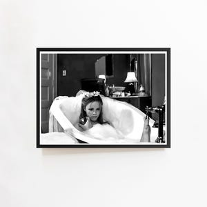 May include: Black and white framed print of a woman in a bathtub. The woman is wearing a floral headpiece and appears to be in a white dress or gown. The bathroom setting includes a mirror, lamp, and faucet.