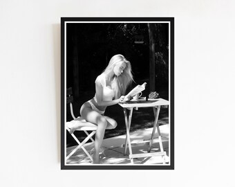 May include: Black and white photograph of a person sitting on a chair and reading a book outdoors. The person is wearing a white top and underwear. A small table with a cup and a plant is next to the person.