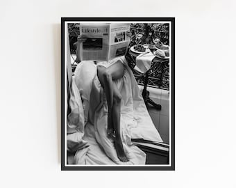 May include: Black and white framed print featuring a person reading a newspaper with the word "Lifestyle" visible. The person is seated on a bed, with legs extended. A small table with food and drinks is in the background.
