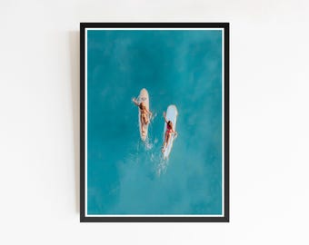 May include: A framed print featuring two people on surfboards in turquoise water. The aerial view shows the individuals lying on their boards, with the ocean's surface creating a vibrant backdrop. The print is encased in a black frame with a white border.