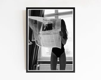 May include: Black and white framed print of a person holding a newspaper in front of their face. The newspaper is open, obscuring the person's face. The person is wearing a black bodysuit. The newspaper's headline is visible.
