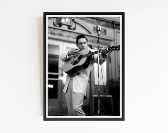 May include: Monochrome framed print featuring a musician playing an acoustic guitar. The musician, in a light-coloured suit, is positioned before a microphone. The image is a medium shot, capturing the musician's performance in a concert setting.
