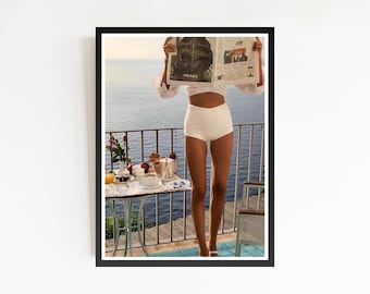 Puede incluir: Una impresión enmarcada que muestra a una persona leyendo un periódico en un balcón con vista al mar. La persona lleva un top blanco y pantalones cortos de cintura alta. Una pequeña mesa está puesta con artículos de desayuno. El periódico dice "PRADA".