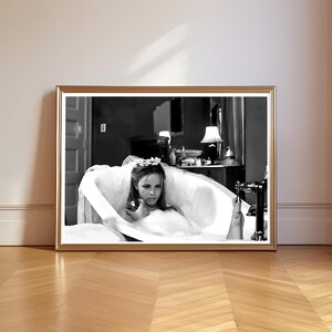 May include: Black and white framed print of a woman in a bathtub. The woman is wearing a veil and a floral crown. The print is in a gold frame and is displayed on a wooden floor.