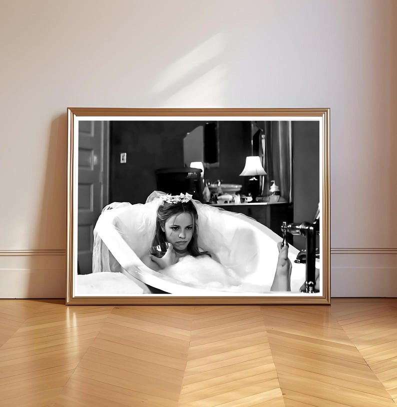May include: Black and white framed print of a woman in a bathtub. The woman is wearing a veil and a floral crown. The print is in a gold frame and is displayed on a wooden floor.