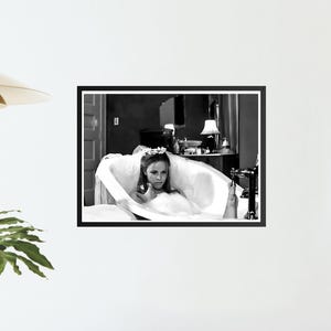 May include: Black and white framed print of a woman in a bathtub. The woman is wearing a floral crown and a white dress. The bathroom has a vintage aesthetic, with a lamp and a mirror in the background.