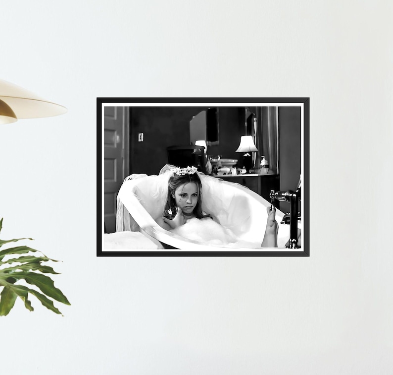 May include: Black and white framed print of a woman in a bathtub. The woman is wearing a floral crown and a white dress. The bathroom has a vintage aesthetic, with a lamp and a mirror in the background.