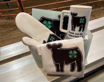 May include: A white kitchen set featuring a mug, an oven mitt, and a towel, all decorated with a black cow silhouette and a green four-leaf clover 4-H emblem. The mug displays the text Thank you for supporting 4-H in black lettering.